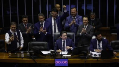 Dez homens vestidos com ternos escuros estão reunidos em plenário. Um homem ao centro está sentado à mesa principal, enquanto os demais ao redor aplaudem e um deles levanta o punho em comemoração. Um cronômetro digital na mesa marca 00:00.