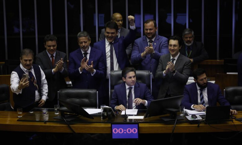 Dez homens vestidos com ternos escuros estão reunidos em plenário. Um homem ao centro está sentado à mesa principal, enquanto os demais ao redor aplaudem e um deles levanta o punho em comemoração. Um cronômetro digital na mesa marca 00:00.
