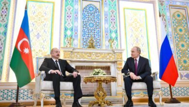 Presidente do Azerbaijão e presidente da Rússia sentados em cadeiras elegantes em sala decorada com padrões geométricos e florais. Bandeiras do Azerbaijão e da Rússia posicionadas ao lado de cada líder, com mesa dourada e arranjo floral entre eles.