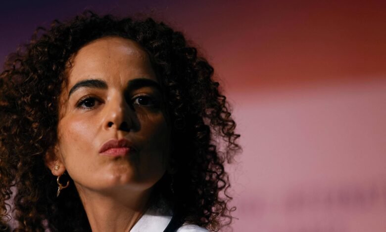 Mulher de pele clara e cabelo cacheado escuro olha diretamente para a câmera com expressão neutra. Ela usa brinco pequeno e camisa branca, com fundo desfocado em tons de vermelho e rosa.