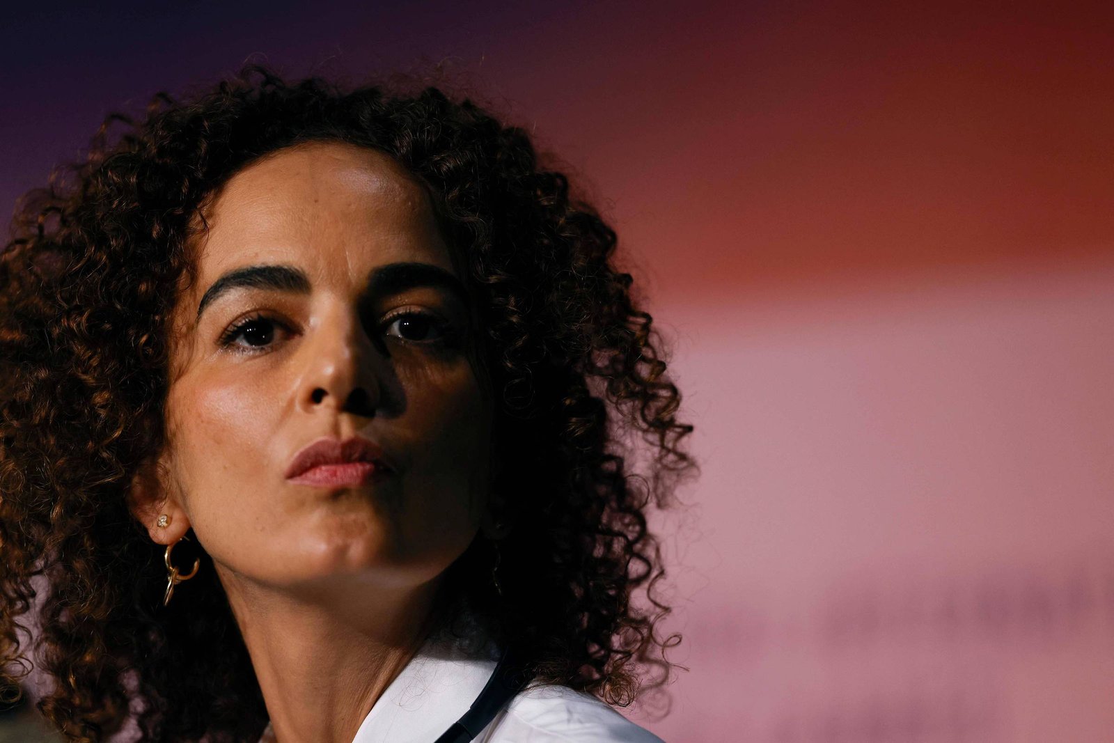 Mulher de pele clara e cabelo cacheado escuro olha diretamente para a câmera com expressão neutra. Ela usa brinco pequeno e camisa branca, com fundo desfocado em tons de vermelho e rosa.