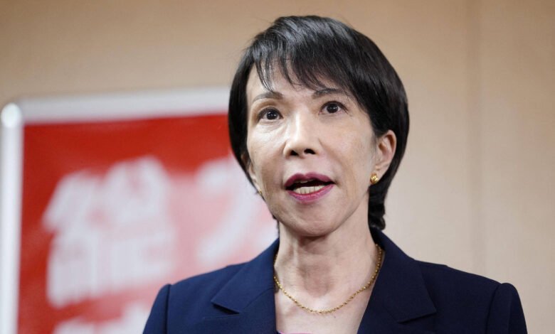 Mulher japonesa em terno azul escuro fala em pronunciamento oficial, com microfones à frente. Fundo desfocado com banner vermelho e branco.