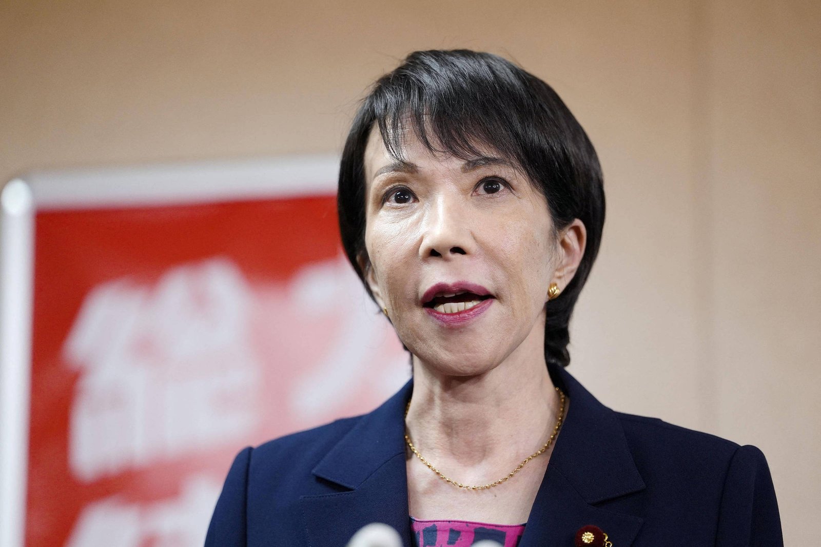 Mulher japonesa em terno azul escuro fala em pronunciamento oficial, com microfones à frente. Fundo desfocado com banner vermelho e branco.