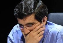 Homem de camisa azul e gravata listrada observa atentamente o tabuleiro de xadrez à sua frente, com a mão direita cobrindo parcialmente o rosto. Peças de madeira claras e escuras estão dispostas no tabuleiro, indicando uma partida em andamento.