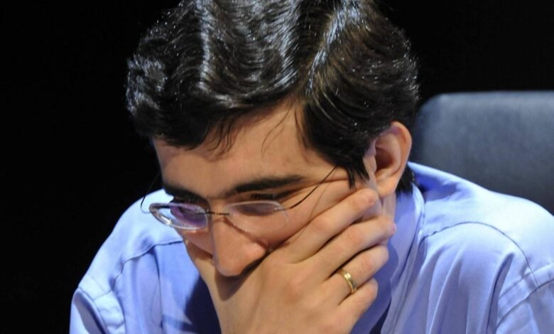 Homem de camisa azul e gravata listrada observa atentamente o tabuleiro de xadrez à sua frente, com a mão direita cobrindo parcialmente o rosto. Peças de madeira claras e escuras estão dispostas no tabuleiro, indicando uma partida em andamento.