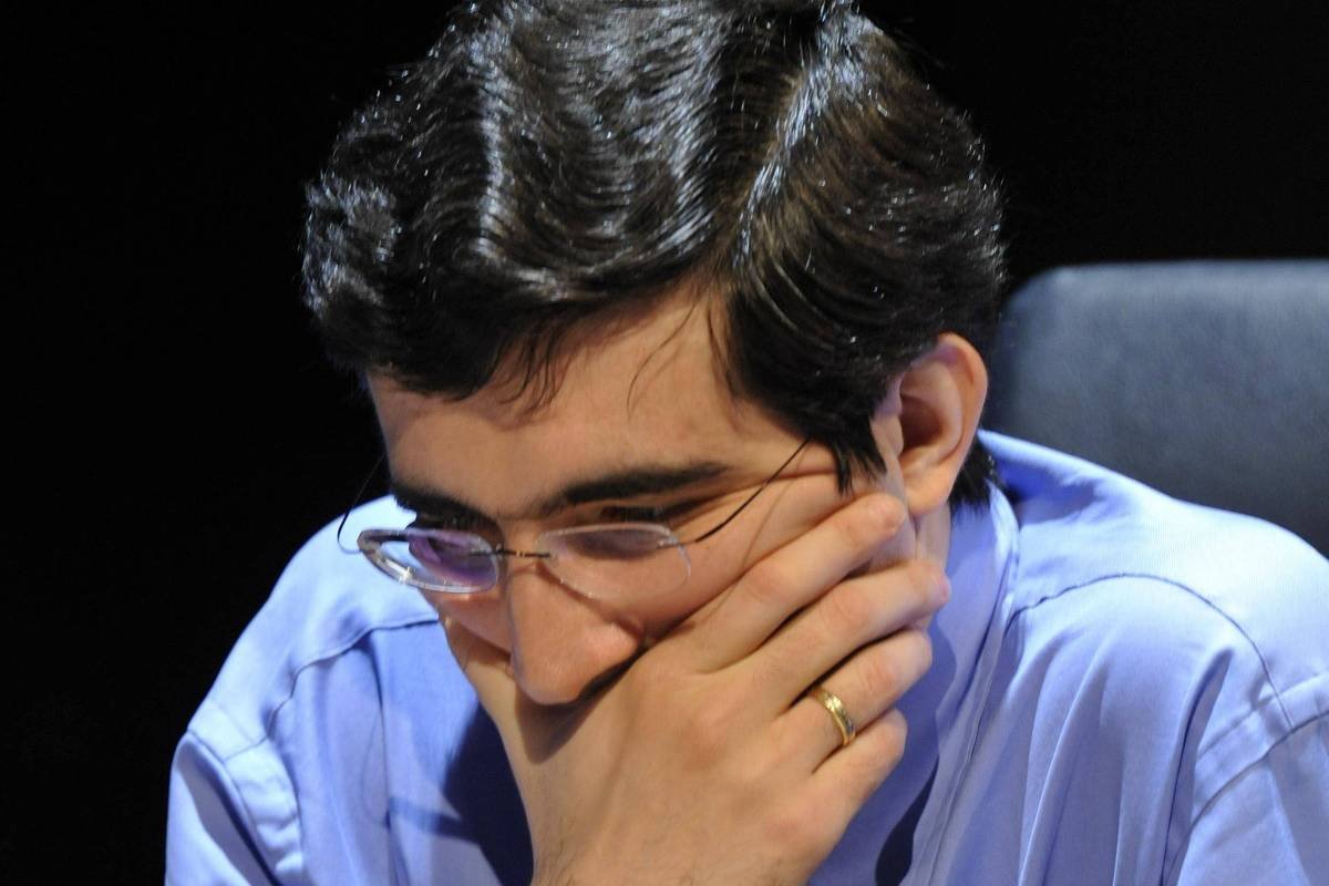 Homem de camisa azul e gravata listrada observa atentamente o tabuleiro de xadrez à sua frente, com a mão direita cobrindo parcialmente o rosto. Peças de madeira claras e escuras estão dispostas no tabuleiro, indicando uma partida em andamento.