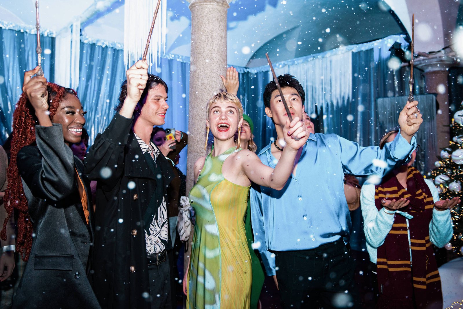 Cinco jovens seguram varinhas mágicas levantadas em ambiente interno decorado com cortinas azuis e neve artificial. Eles sorriem e olham para cima, com uma árvore de Natal ao fundo à direita.