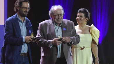 Quatro pessoas estão alinhadas no palco durante cerimônia. Dois homens no centro seguram troféus, um deles usa blazer cinza e o outro, blazer azul. Homem à esquerda veste terno cinza e gravata azul. Mulher à direita usa vestido amarelo claro com detalhes nos ombros. Fundo escuro com iluminação roxa.