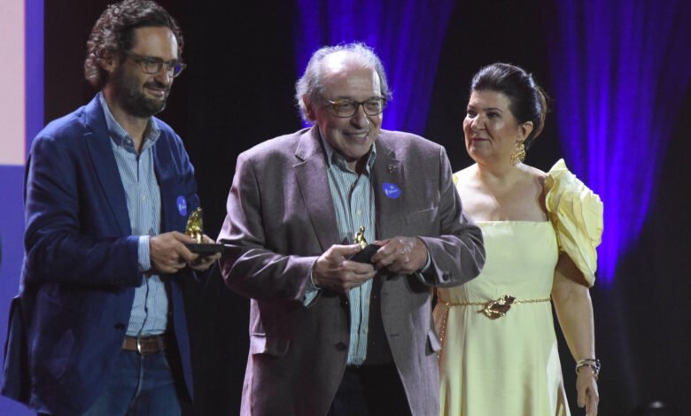 Quatro pessoas estão alinhadas no palco durante cerimônia. Dois homens no centro seguram troféus, um deles usa blazer cinza e o outro, blazer azul. Homem à esquerda veste terno cinza e gravata azul. Mulher à direita usa vestido amarelo claro com detalhes nos ombros. Fundo escuro com iluminação roxa.