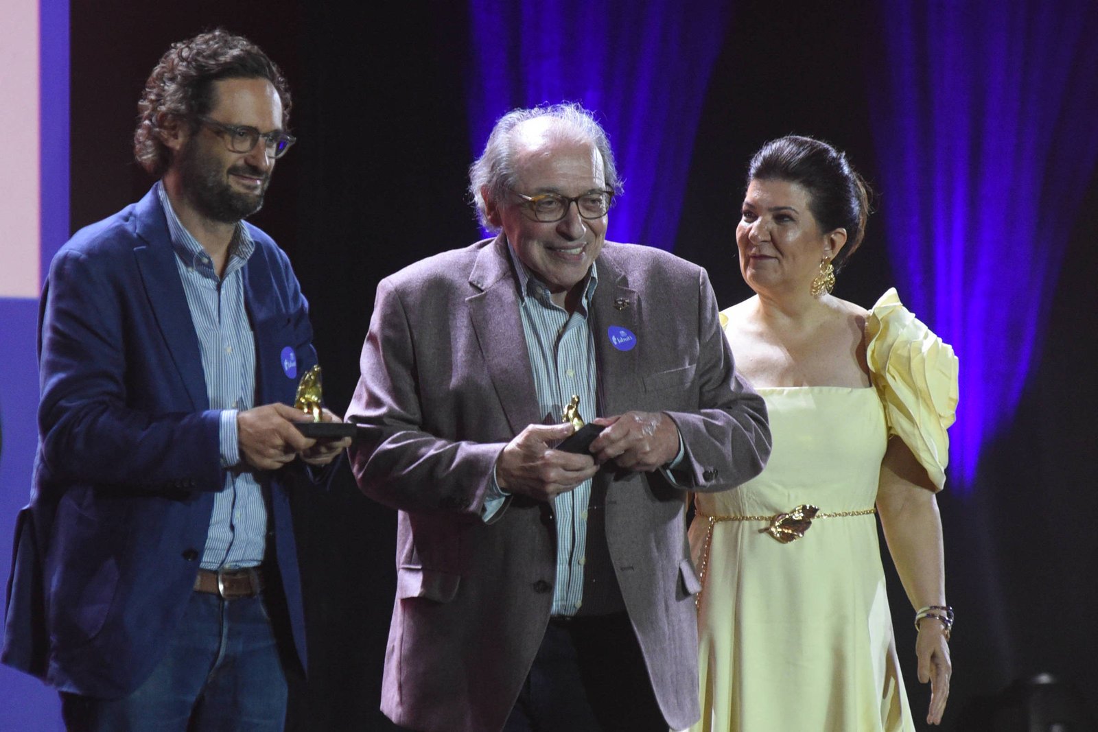Quatro pessoas estão alinhadas no palco durante cerimônia. Dois homens no centro seguram troféus, um deles usa blazer cinza e o outro, blazer azul. Homem à esquerda veste terno cinza e gravata azul. Mulher à direita usa vestido amarelo claro com detalhes nos ombros. Fundo escuro com iluminação roxa.