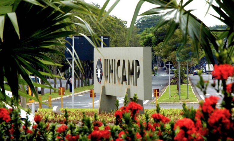 A imagem mostra a entrada da Universidade Estadual de Campinas (UNICAMP), com um letreiro em destaque que exibe o nome da instituição. Ao redor, há vegetação exuberante, incluindo palmeiras e flores vermelhas. Ao fundo, é possível ver uma via com veículos e sinalização.