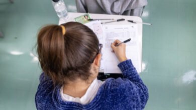 A imagem mostra a parte de trás de uma estudante com cabelo preso em um coque, usando um suéter azul. Ela está escrevendo em uma folha de papel sobre uma mesa, que também contém uma garrafa de água, canetas e outros papéis. O ambiente parece ser uma sala de aula, com piso verde.