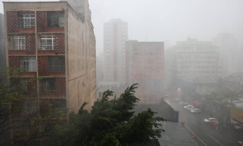 A imagem mostra um cenário urbano durante uma forte chuva. Vários prédios são visíveis, com um deles em primeiro plano apresentando janelas e uma fachada de tijolos. A visibilidade é baixa devido à intensidade da chuva, e árvores balançam com o vento. O ambiente parece molhado e cinzento, refletindo as condições climáticas adversas.