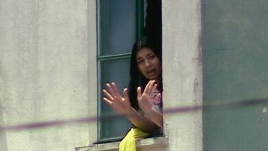 Mulher com cabelo preto aparece na janela aberta de um prédio claro, com as mãos levantadas em sinal de parada ou comunicação. Ela veste roupa clara e há um tecido amarelo pendurado no parapeito da janela.