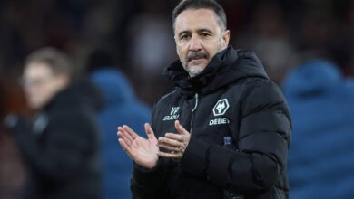 Treinador Vítor Pereira com casaco preto acolchoado com logotipo do Wolverhampton Wanderers aplaude em campo. Fundo desfocado mostra outras pessoas e ambiente de estádio à noite.