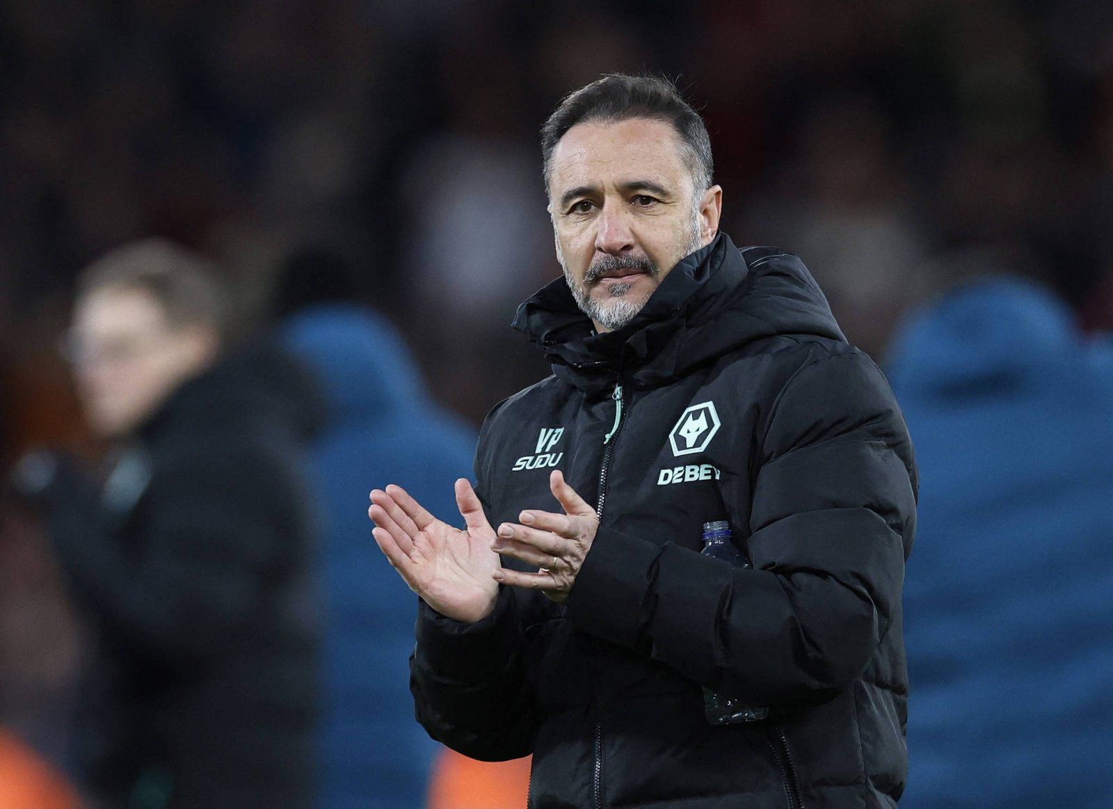 Treinador Vítor Pereira com casaco preto acolchoado com logotipo do Wolverhampton Wanderers aplaude em campo. Fundo desfocado mostra outras pessoas e ambiente de estádio à noite.
