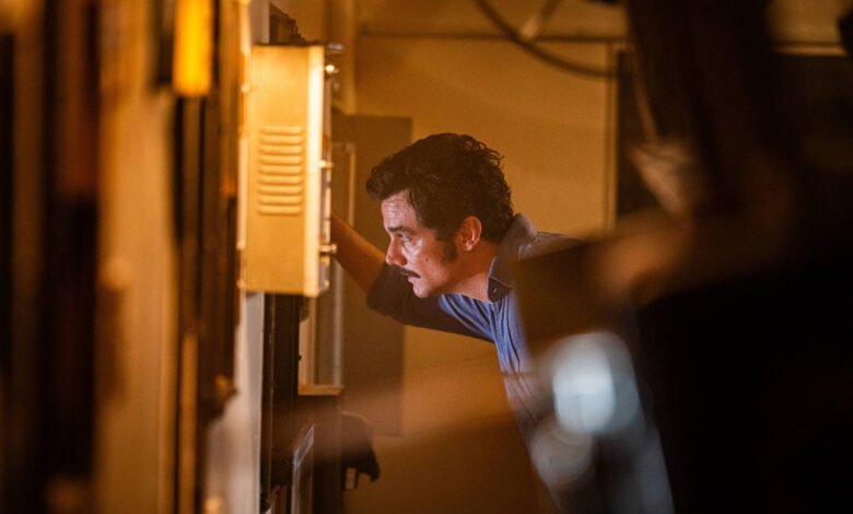 Homem de camiseta azul inclina-se para examinar um painel elétrico aberto em ambiente interno com iluminação amarelada. O foco está no rosto concentrado e na mão que toca o painel.