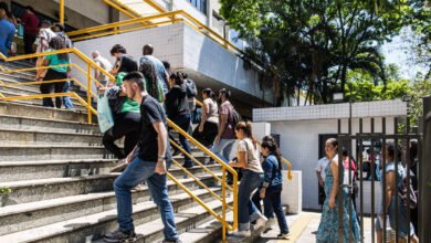 Grupo de pessoas subindo escada externa com corrimão amarelo em prédio branco. Ambiente ao ar livre com árvores e sol forte. Algumas pessoas carregam mochilas e bolsas.
