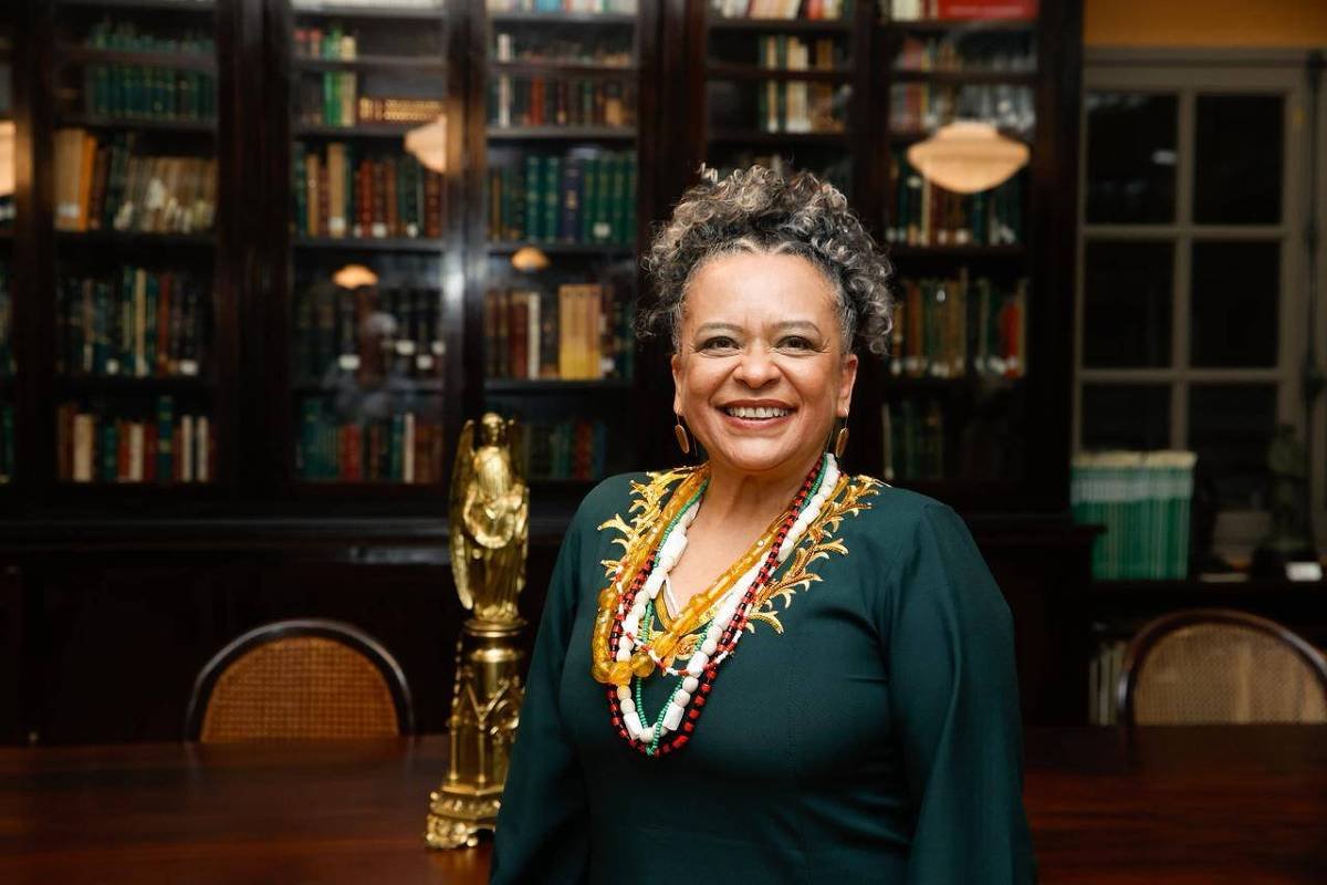 Mulher de cabelos grisalhos encaracolados sorri em biblioteca com estantes cheias de livros ao fundo. Ela veste blusa verde escura e vários colares coloridos. Mesa de madeira e cadeira com encosto de palha aparecem à frente.