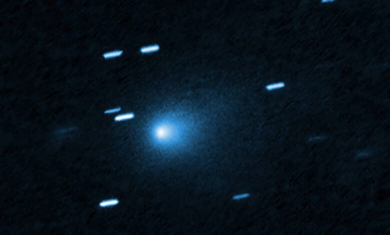 Cometa azul brilhante no centro da imagem com uma cauda difusa ao redor. Traços de estrelas aparecem como linhas claras ao redor, indicando movimento ou longa exposição.