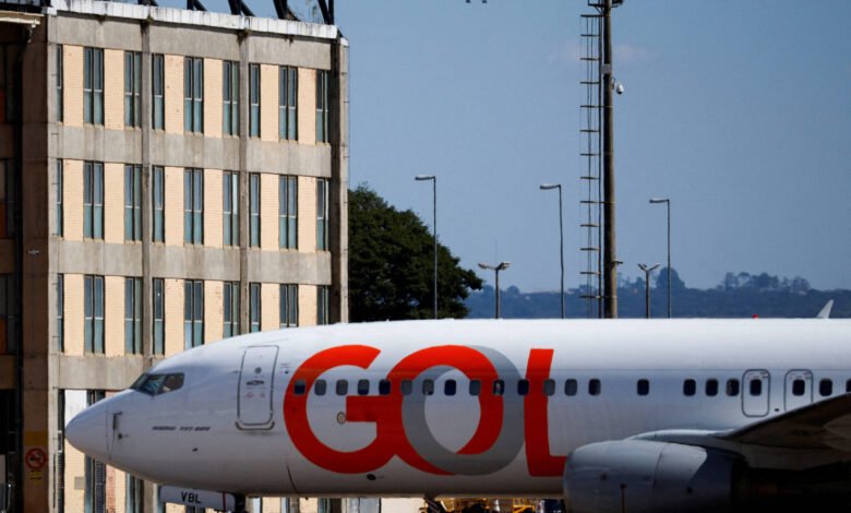Avião da Gol estacionado em primeiro plano próximo a um prédio de janelas retangulares. Ao fundo, outro avião está em aproximação para pouso sob céu azul.