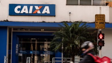 Fachada da agência da Caixa Econômica Federal com letreiro azul e branco. À direita, semáforo de pedestres vermelho e placa amarela indicando cuidado com ônibus. Pessoa com capacete e mochila vermelha passa em primeiro plano.