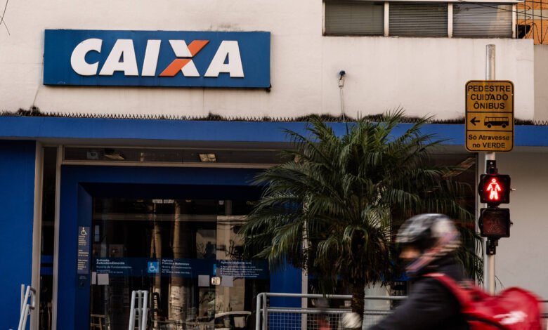 Fachada da agência da Caixa Econômica Federal com letreiro azul e branco. À direita, semáforo de pedestres vermelho e placa amarela indicando cuidado com ônibus. Pessoa com capacete e mochila vermelha passa em primeiro plano.