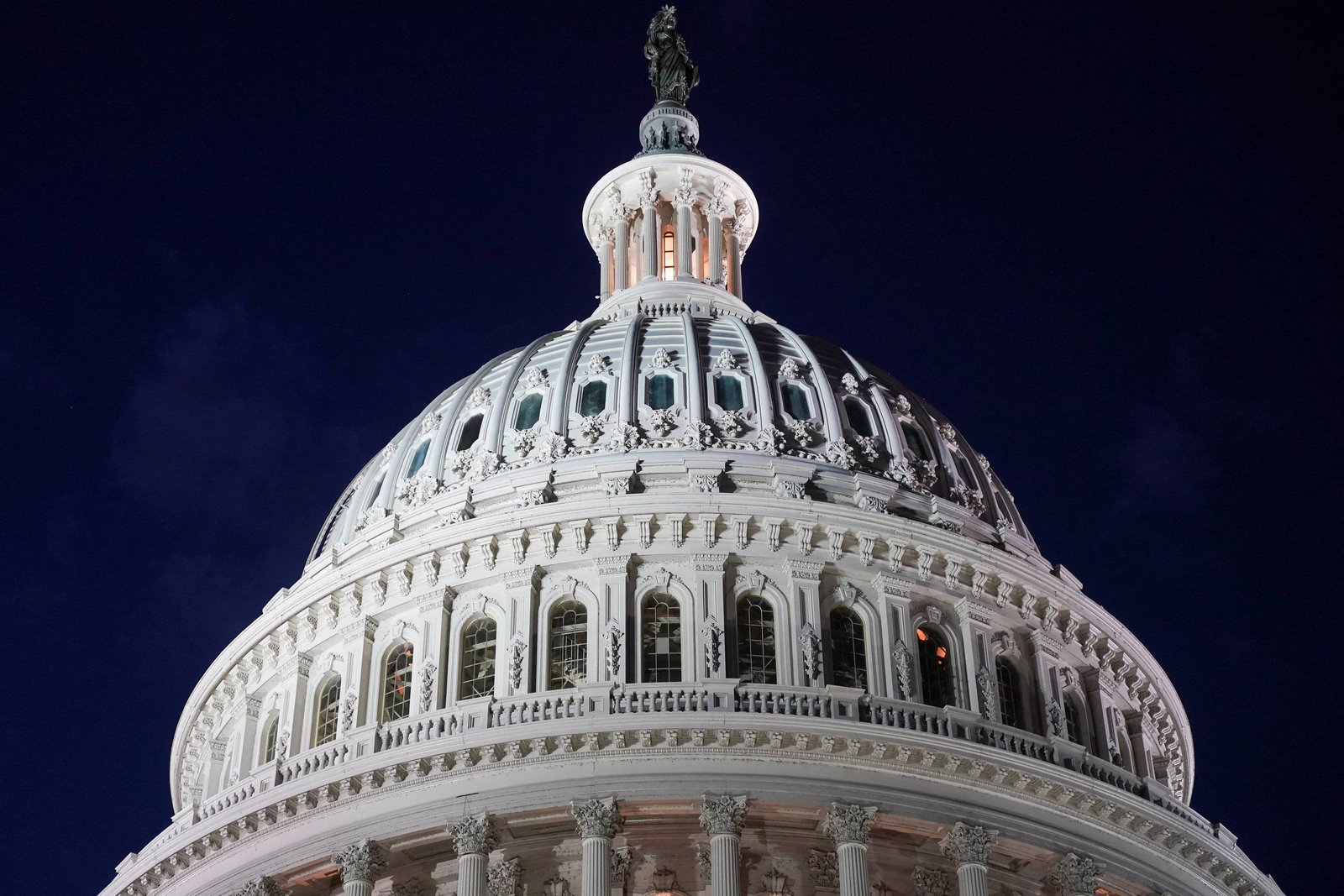 Vista frontal da cúpula branca do Capitólio dos Estados Unidos iluminada contra o céu noturno escuro. Detalhes arquitetônicos e janelas são visíveis.