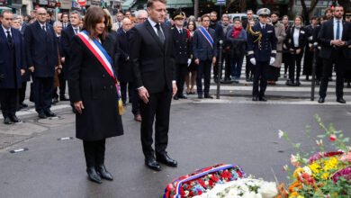 Dois líderes em trajes formais, um com faixa tricolor, estão em posição de respeito diante de coroas de flores na rua. Atrás deles, grupo de pessoas, incluindo oficiais fardados e civis, observa a cerimônia em ambiente urbano.