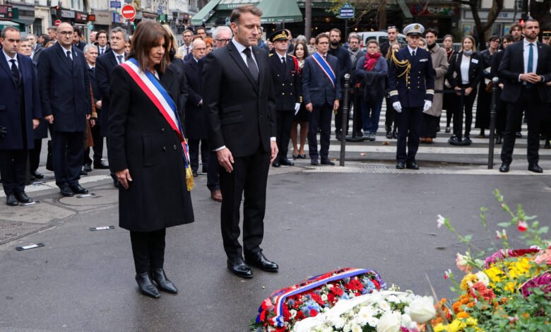 Dois líderes em trajes formais, um com faixa tricolor, estão em posição de respeito diante de coroas de flores na rua. Atrás deles, grupo de pessoas, incluindo oficiais fardados e civis, observa a cerimônia em ambiente urbano.