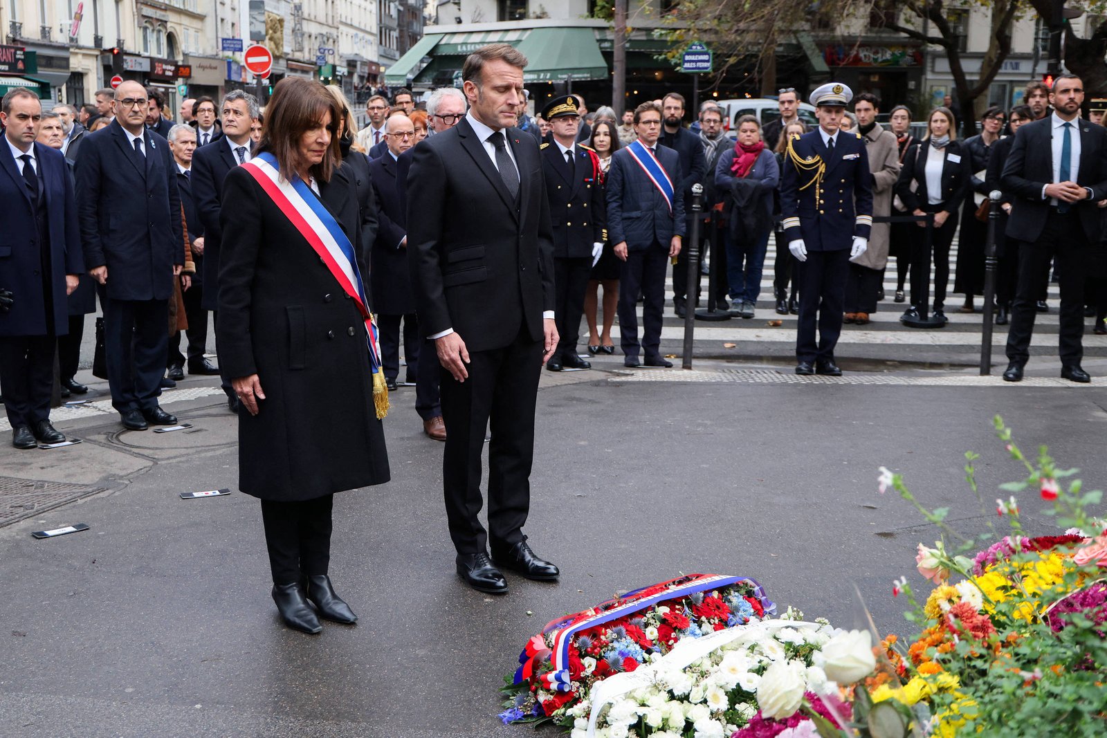 Dois líderes em trajes formais, um com faixa tricolor, estão em posição de respeito diante de coroas de flores na rua. Atrás deles, grupo de pessoas, incluindo oficiais fardados e civis, observa a cerimônia em ambiente urbano.