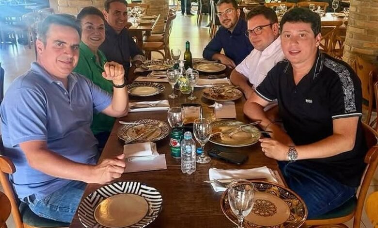 Seis homens sentados ao redor de uma mesa de madeira em restaurante com decoração rústica, incluindo pilares de tijolos e teto com estrutura de madeira. A mesa está posta com pratos, talheres, copos e garrafas de água. Ao fundo, outras pessoas estão em pé e sentadas, com luz natural entrando pelas janelas.