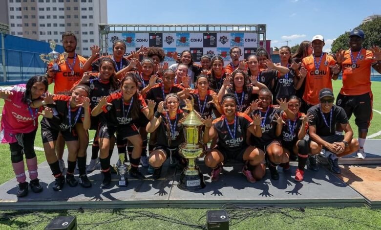 Jogadores disputam a bola em campo durante a final da Primeira Copa CDHU de Futebol na Zona Leste. O evento atraiu grande público ao longo de dois meses de competição e elegeu campeões regionais.